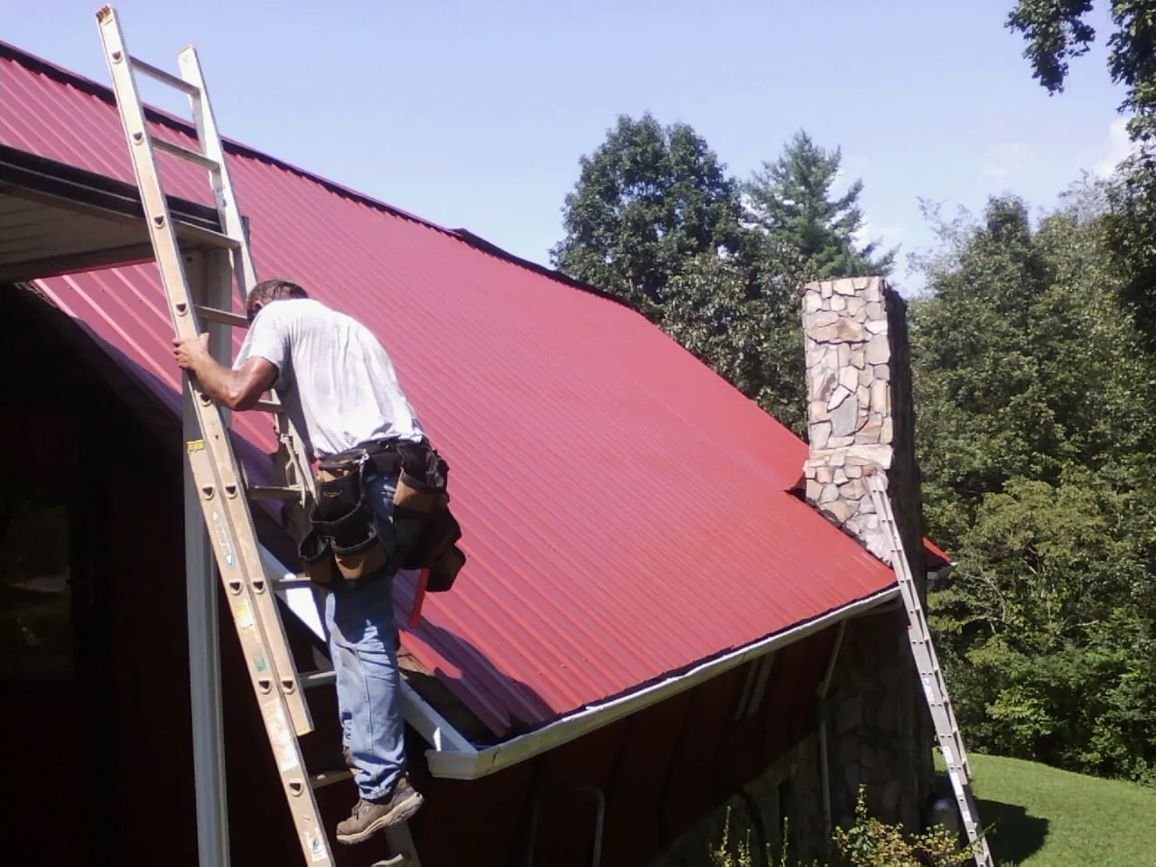 Roofer on a ladder beside a red metal roof and stone chimney — Soffit & Fascia Repair by Roofing Brothers in Dravosburg
